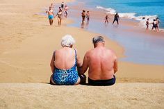 an older couple sitting on the beach looking out at the water and people swimming in the ocean