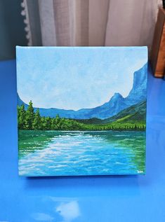 a painting of a lake with mountains in the background on a blue tablecloth next to a window