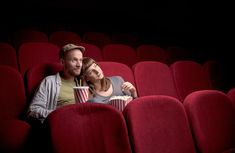 a man and woman sitting in the middle of a movie theater with red seats holding drinks