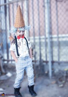 a little boy dressed up as a clown with a hat and suspenders, standing in front of a fence