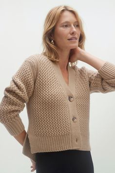 a woman wearing a tan cardigan sweater and black skirt with her hand on her chin