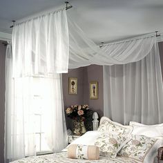 a white bed sitting next to a window covered in sheer curtains and pink flowers on top of it