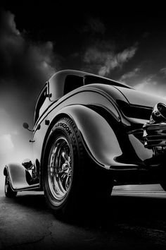 a black and white photo of an old fashioned car in the dark with clouds behind it