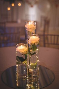 three white roses are placed in glass vases on a table with lights around them