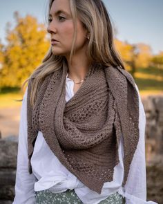 a woman wearing a brown knitted shawl and white shirt with trees in the background