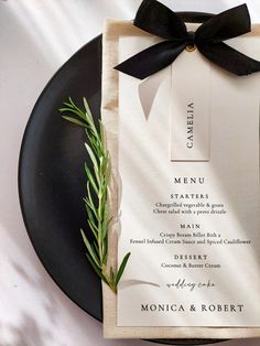 a black plate topped with a menu on top of a white table cloth covered in greenery