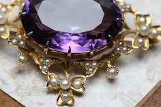 "A beautiful antique Late Victorian to Edwardian 14k yellow gold large amethyst and seed pearl brooch. A lovely design with shamrocks at the four corners and the gold work set with 44 tiny seed pearls. The brooch measures 1 5/8\" x 1 1/2\". There is a large oval faceted amethyst in the center prong set and measures approx. 21mm x 15mm x 10mm (16.7ct). Set down into the gold are 44 small seed pearls with the size ranging around .75mm to 2.5mm . The back is fitted with a ball hinge and a locking c Elegant Purple Gemstone Brooches, Elegant Amethyst Brooches For Wedding, Elegant Purple Brooches For Anniversary, Elegant Gold Amethyst Brooches, Purple Victorian Wedding Brooches, Gold Work, Pearl Brooch, Seed Pearl, Four Corners