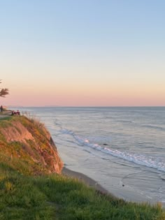 people are sitting on the edge of a cliff overlooking the water and beach at sunset