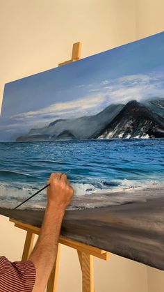 a man is painting on an easel by the ocean with mountains in the background