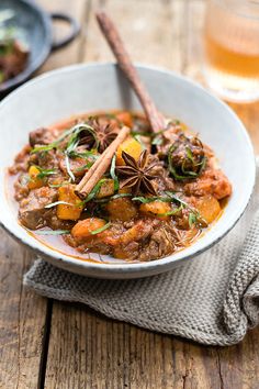 a white bowl filled with stew and spices on top of a wooden table next to a glass of beer