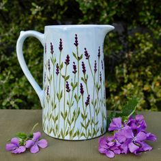 purple flowers are sitting on a table next to a white pitcher with green and purple designs