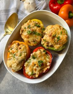 three stuffed peppers in a white bowl next to some tomatoes and bell peppers on a table