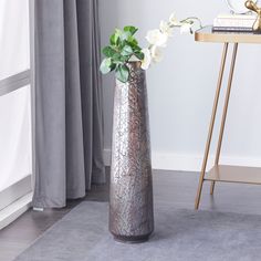 a silver vase with flowers in it sitting on a rug next to a table and window