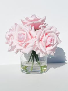 three pink roses in a clear vase on a white surface with shadows from the wall behind them