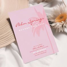 a pink card with palm trees on it sitting next to a hat and some flowers