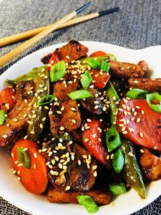 a white plate topped with meat and veggies covered in sesame seeds next to chopsticks