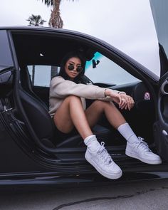 a woman sitting in the door of a car with her legs crossed and wearing white sneakers