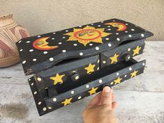 a hand is holding an ornate wooden box with sun and moon designs on the lid