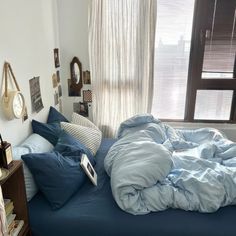 an unmade bed with blue sheets and pillows in a bedroom next to a window