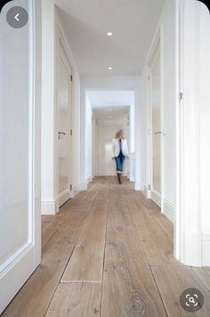 a woman walking down a long hallway with white walls and wood flooring on both sides