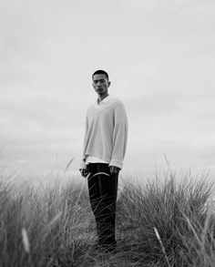 a black and white photo of a man standing in tall grass with his hands in his pockets