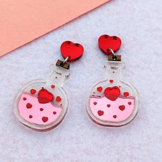 These love potions are perfect for Valentine's Day. They feature red hearts bottled in a pink elixir. There are red engraved details surrounding pink and red shiny acrylic, inside of a clear bottle with wooden corks on top. They are shiny and reflective to add a little sparkle to your life. Dimensions: 1.5 in (38 mm) L x 1.0 in (26 mm) W The drop length is about 2.0 in (51 mm). Materials: Mirror Acrylic Hypoallergenic, Nickel Free, Stainless Steel Shipping: All of our earrings are made-to-order to cut down on waste. Your earrings will be produced and shipped within a week after you order. Care: Do not soak earrings in water. Materials are water resistant, but it is not recommended that you swim, shower, or bath with your earrings on. Avoid getting lotions, oils, and perfumes on your earrin Red Heart Earrings For Gift, Red Round Heart Earrings For Gift, Love Potions, Bottle Cute, Valentine Earrings, Clear Bottle, Valentine Jewelry, Clay Dangle Earrings, Valentines Earrings