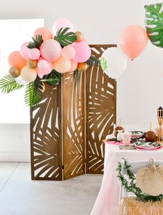 a table topped with lots of balloons and greenery next to a wooden screen divider