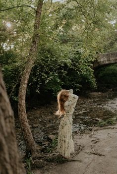 a woman standing in front of a tree next to a river wearing a white dress