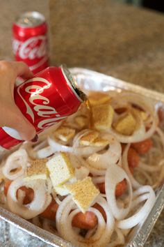 a person is pouring coke into a tray of food with onions and hot dogs on it