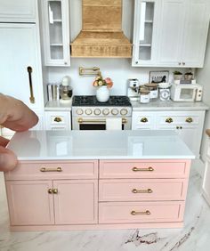 a pink kitchen with white cabinets and gold handles on the countertop is being held up by someone's hand