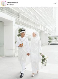 two people dressed in white holding hands and walking down the street with flowers on their hand