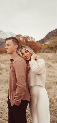 a man and woman are standing together in the grass with mountains in the back ground