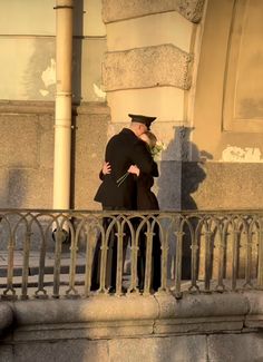 two men in black coats embracing each other on a balcony with wrought iron railings