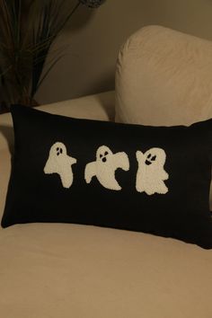 a black pillow with three white ghostes on it sitting on a couch next to a potted plant