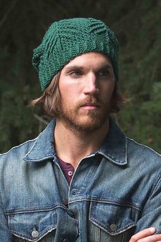 a man with long hair wearing a green knitted hat and denim jacket standing in front of trees