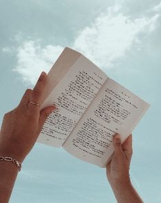 two hands hold open a book in front of a blue sky with white fluffy clouds