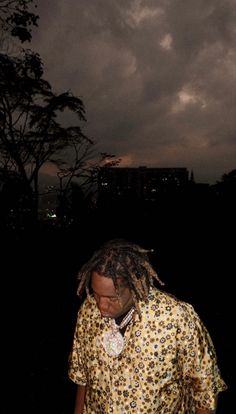 a man with dreadlocks standing in front of a dark sky at night wearing a yellow and black shirt