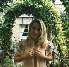 a woman standing in front of a green archway with plants around her and holding hands together