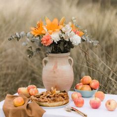 a table topped with a vase filled with flowers next to apples and pies on top of a white table cloth