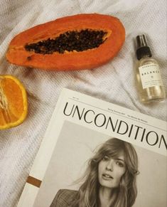 the magazine is next to an orange and some other items on a white sheet with a bottle of cologne