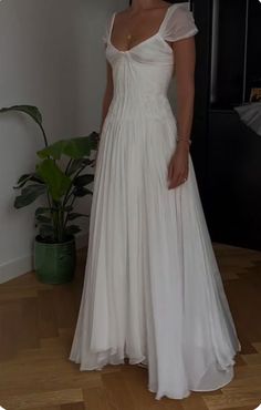 a woman in a white dress standing on a wooden floor next to a potted plant