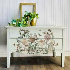 a white dresser with flowers painted on the front and sides, next to a gold vase