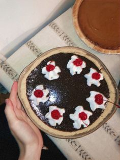 a person holding a chocolate cake with cherries on it and another pie in the background