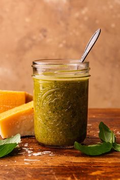 a jar filled with green pesto next to slices of cheese and a silver spoon