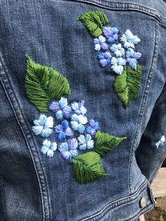 the back of a woman's jean jacket with blue flowers embroidered on it and green leaves