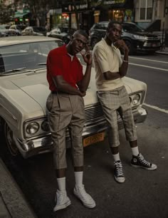 two young men sitting on the hood of a car in front of a city street