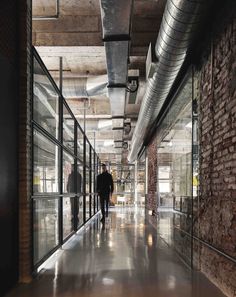 a man walking down a long hallway in an office building with glass walls and exposed pipes