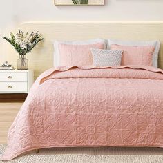 a bed with pink bedspread and pillows in a white room next to a plant