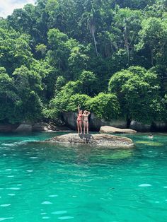 two people standing on a rock in the middle of a body of water surrounded by trees