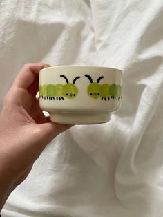 a hand holding a white bowl with green bugs painted on the side and black eyes